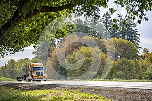 Yellow big rig bonnet semi truck with step down semi trailer running on the green highway road