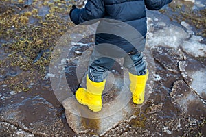 bright yellow rubber boots on the feet of a boy walking through mud, snow and puddles in early spring