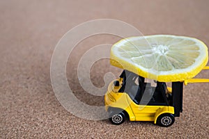 A bright yellow toy forklift carries a slice of lemon. Close-up miniatures on balsa wood background. Modeling