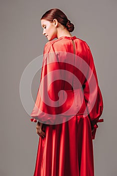 Bright young lady in red dress with long wide sleeves and sexy neckline on high heels mooving on grey studio background
