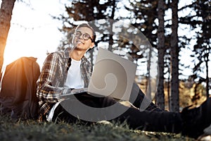 Bright young male student in casual outfit using notebook or laptop while sitting on the grass at the park