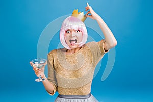 Brightful party time of funny young woman with champagne, golden crown on head having fun on blue background. Cut pink