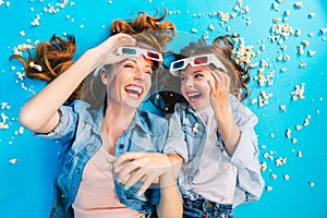 Brightful stylish image from above excited mother and daughter laying on blue floor in popcorn, laughing in 3D glasses