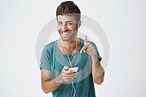Brightly smiling unshaved spanish guy in blue t-shirt, holding smartphone, listening music with headphones, laughing and