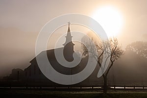 Brilliant bright sun silhouettes small church and tree in fog at dawn photo