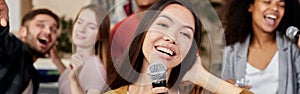 Bring joy. Young pretty asian woman holding microphone and singing while having fun with friends at home. Group of