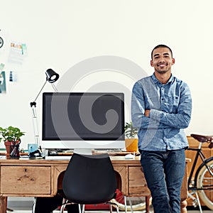 He brings designs to life. Portrait of a casually-dressed young man sitting on his desk in an office.