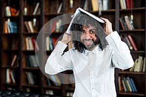 A broadly smiling Egyptian student holds a book above his head in the library against the background of books