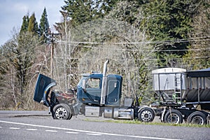 Broken big rig semi truck with an open hood and loaded semi trailer stands on the side of the road waiting for a mobile repair