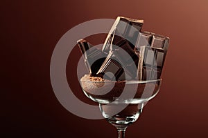 Broken chocolate bar in glass with cocoa powder on a brown background