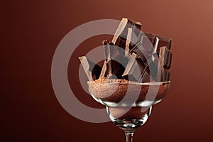 Broken chocolate bar in glass with cocoa powder on a brown background