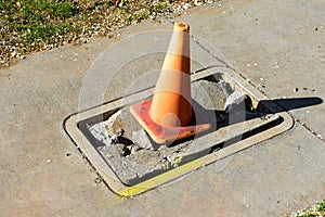 Broken concrete utility box cover plate on the sidewalk marked with orange traffic cone for visibility and safety