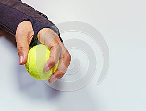 Broken hand in black orthosis holding a yellow tennis ball for training and recovery on light gray background