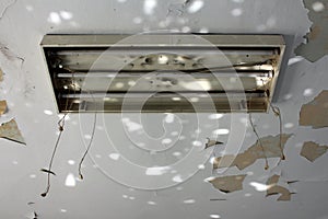 Broken overhead fluorescent lamp fixture with three fluorescent lamps and hanging disconnected electrical wires on cracked ceiling