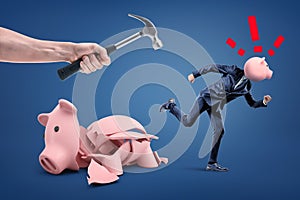 Broken piggy bank and businessman with piggy bank head running from hand isolated with hammer on blue background