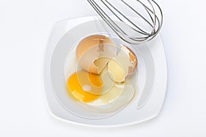 Broken raw egg and wire stirrer in a porcelain dish on white