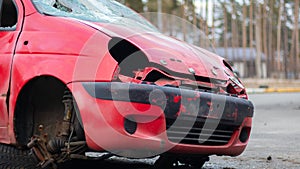 A broken, rumpled small red car stands on the side of the road without headlights and a wheel after a powerful blow