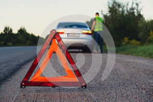 Broken silver luxury car emergency accident. Man driver installing red triangle stop sign on road. Sport automobile