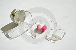 A broken small glass bottle with metal cap, two pink heart-shaped tablets fell out of it, on dirty white background