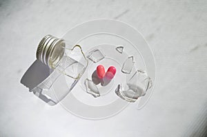 A broken small glass bottle with metal cap, two pink heart-shaped tablets fell out, on dirty white background, side view