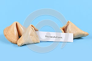 Broken up and whole fortune cookie with motivational text `This too shall pass` on paper note on blue background
