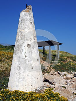 Broken white conical concrete structure next to a shelter on the coastal cliffs surrounded by yellow spring flowers and a bright