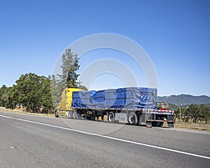 Broken yellow big rig semi truck with loaded flat bed semi trailer sits on the side of a highway waiting for a mobile repair crew