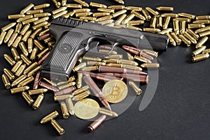 Bronze and brass gun bullets scattered on dark table, black pistol, golden bitcoin coins near - illegal use of cryptocurrency to