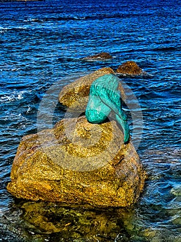 Bronze mermaid sculpture called Sirenella di L`Isula Rossa sitting on a rock in the Mediterranean Sea