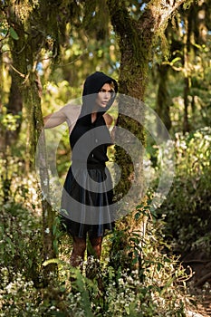 Brooding dreamy young girl blonde, black dress with a hood standing by a tree