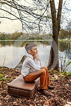 A brooding rustic simpleton fellow sitting on a retro old-fashioned suitcase on the bank of a river lake