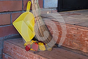 Broom with a red handle and a yellow dustpan, propped against a brick wall next to a pile of autumn leaves on a wooden brown porch