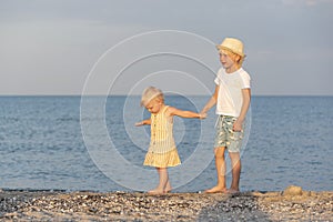 Brother holds his little sister by hand and walks barefoot along the beach