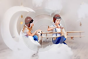 Brother and sister in aviator helmet playing with big wooden airplane in white clouds. Children and their dog together in sky with