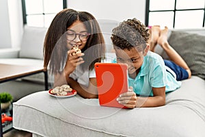 Brother and sister eating cookies using touchpad lying on sofa at home