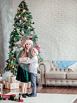 Brother and sister give and unpack gifts under the Christmas tree, sitting by the fireplace, on Christmas morning