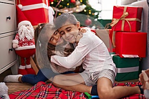 Brother and sister hugging each other sitting on floor by christmas gifts at home