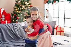 Brother and sister hugging each other standing by christmas tree at home