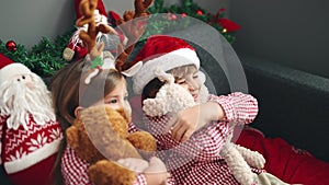 Brother and sister hugging teddy bears sitting on sofa by christmas decoration at home