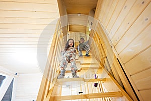Brother with sister in pajama sit on stairs in cozy wooden tiny cabin house. Life in countryside