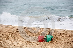 Brother and sister playing with sand and water on a tropical beach, dressed in protective wetsuit