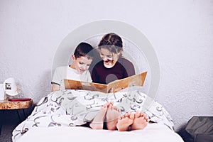 Brother and sister read a book together. Happy and joyful children with bare feet on the bed. The legs are covered with a blanket.