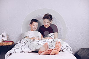 Brother and sister read a book together and laugh. Happy and joyful children with bare feet on the bed. The legs are covered with