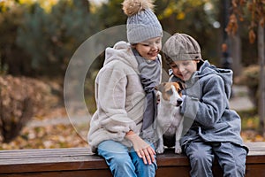 Brother and sister sit in an embrace with a dog on a bench for a walk in the autumn park. Boy, girl and jack russell