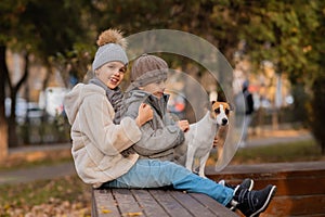 Brother and sister sit in an embrace with a dog on a bench for a walk in the autumn park. Boy, girl and jack russell