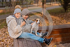 Brother and sister sit in an embrace with a dog on a bench for a walk in the autumn park. Boy, girl and jack russell