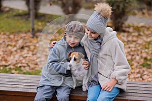 Brother and sister sit in an embrace with a dog on a bench for a walk in the autumn park. Boy, girl and jack russell