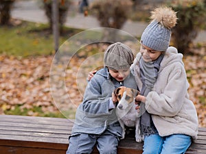 Brother and sister sit in an embrace with a dog on a bench for a walk in the autumn park. Boy, girl and jack russell