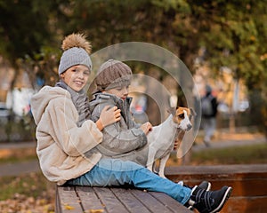 Brother and sister sit in an embrace with a dog on a bench for a walk in the autumn park. Boy, girl and jack russell