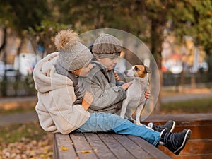 Brother and sister sit in an embrace with a dog on a bench for a walk in the autumn park. Boy, girl and jack russell